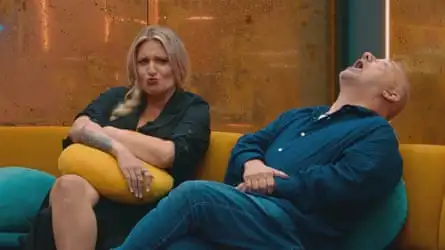 A white woman scrunches up her faces as she looks forward sat on a sofa, the white male to her right leaning back as if howling
