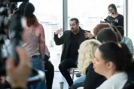A white man with a beard and moustache sits on a chair and gesticulates