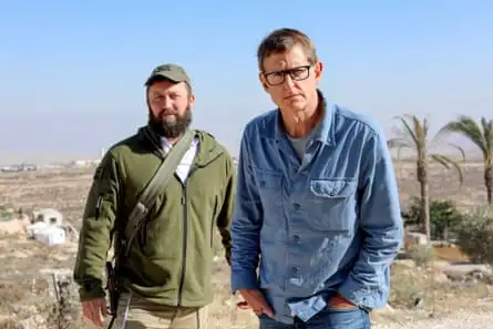 A beared white man in a cap stands looking ahead behind a white man with a denim shirt and dark rimmed glasses looking at the viewer