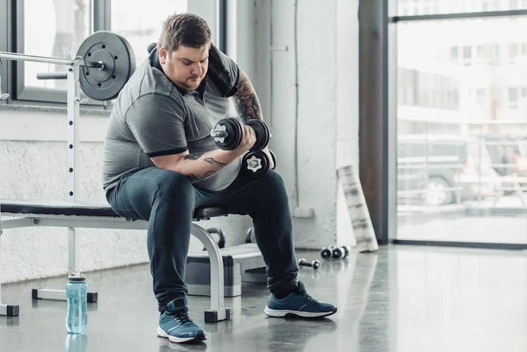 An overweight man with tattoos on his arms performs a barbell curl at the gym while seated on a bench.