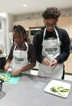 Young people wearing aprons chopping cucumbers in a kitchen.