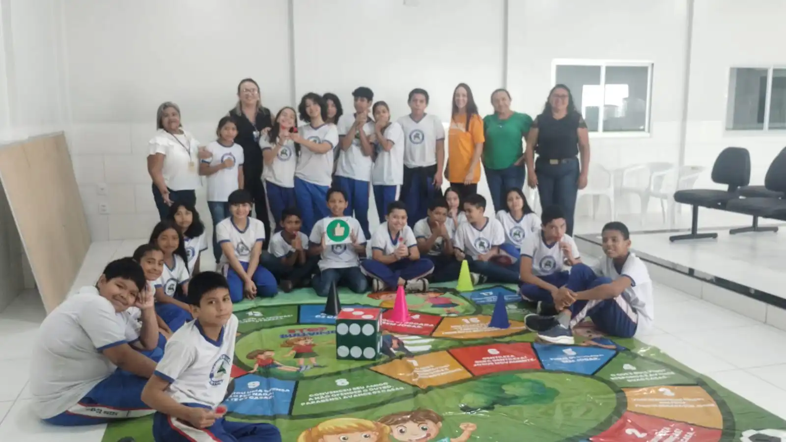A imagem colorida mostra um grupo de cerca de 15 pessoas, incluindo adultos e jovens, participando de uma atividade em um espaço coberto. A maioria dos jovens veste uniformes escolares, e alguns adultos parecem ser educadores. As pessoas estão em círculo, algumas com os braços estendidos e segurando canetas, sugerindo um jogo ou exercício de equipe. Há uma parede clara com um padrão geométrico decorativo e, ao fundo, árvores e um carro.