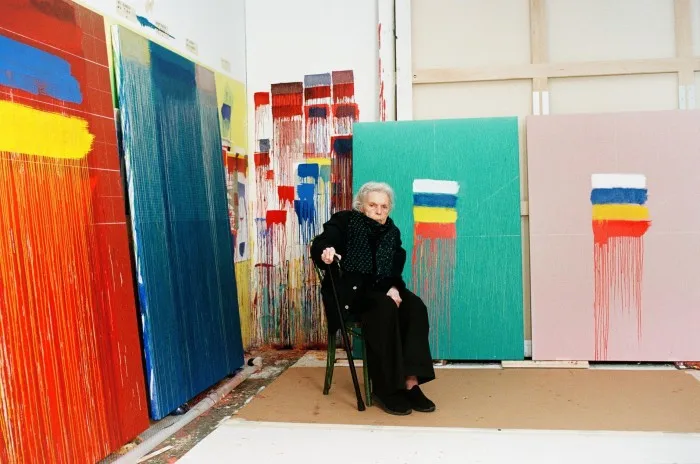 An elderly woman sits in an artist’s studio surrounded by colourful abstract artworks; she holds a walking stick and is swaddled in a coat and scarf