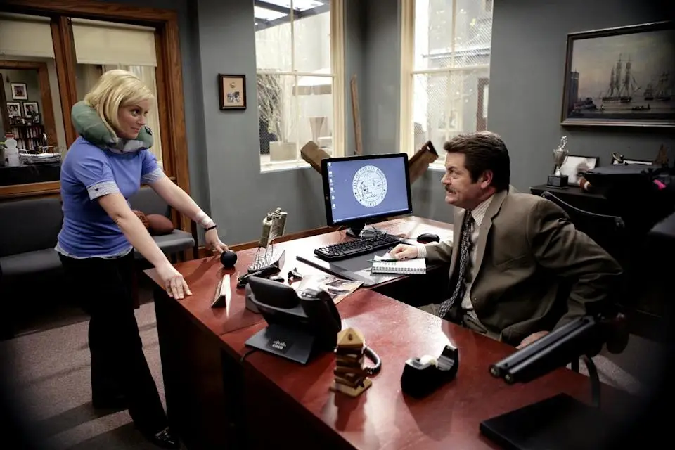 A woman wearing a neck pillow stands at the desk of a seated man