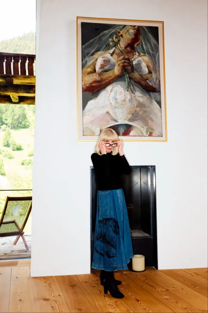 Woman in black-framed glasses, with a silver bob, black sweater and a long denim skirt stands in front of an expressionistic oil painting of a woman carrying white roses, wearing in a white petticoat with suspenders, being viewed from below.  