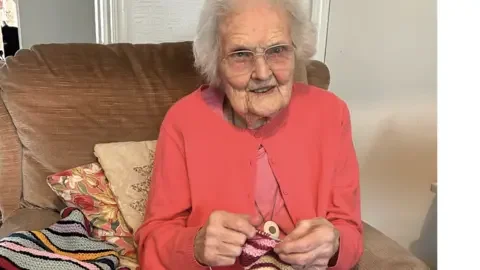 Cat Protection Beryl Jenkins is facing the camera with her knitting equipment in her hand. She has glasses on and a pink cardigan, sitting on a sofa.