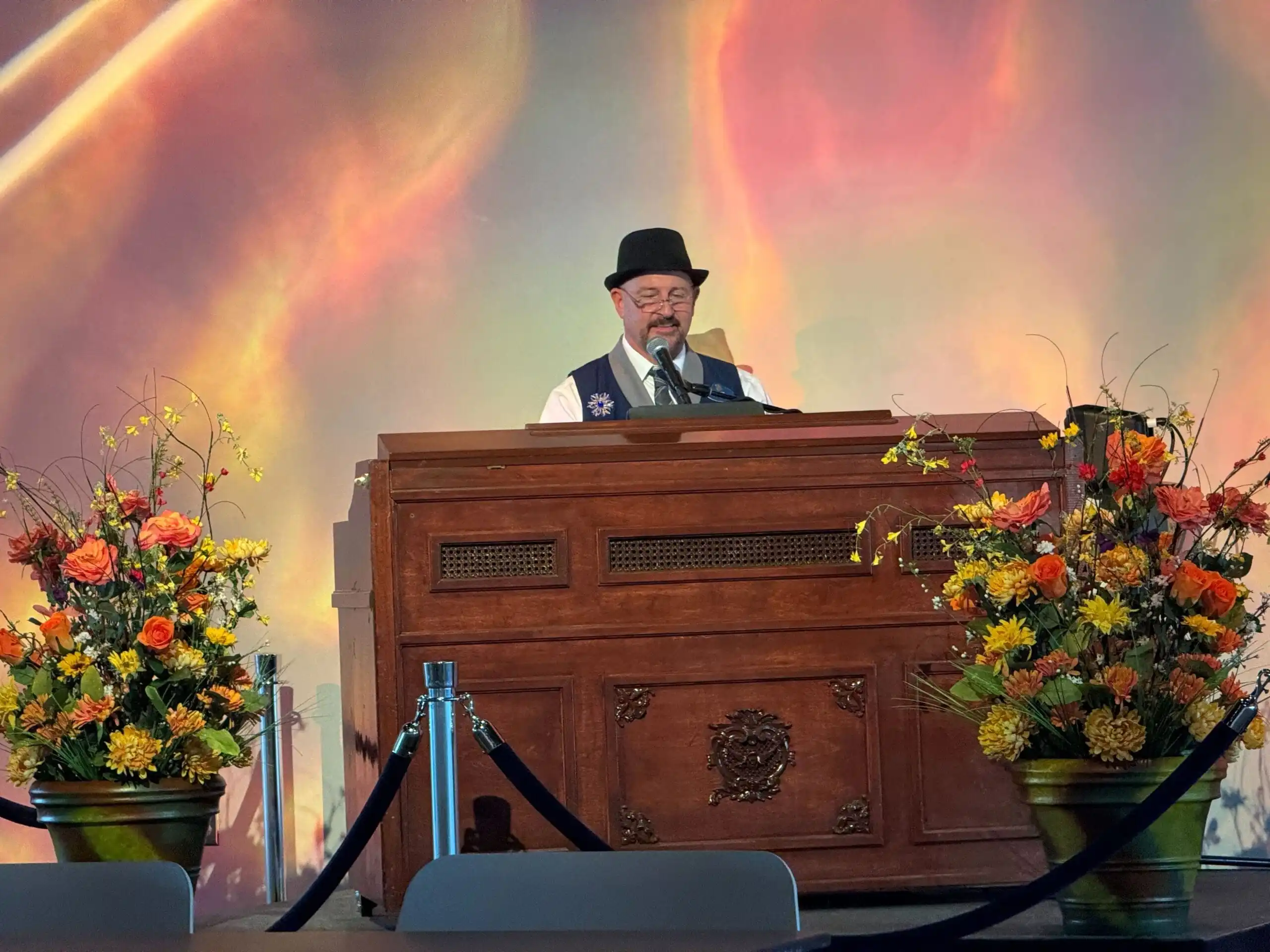 A man in a bowler hat plays a large organ onstage at EPCOT, flanked by two large floral arrangements.