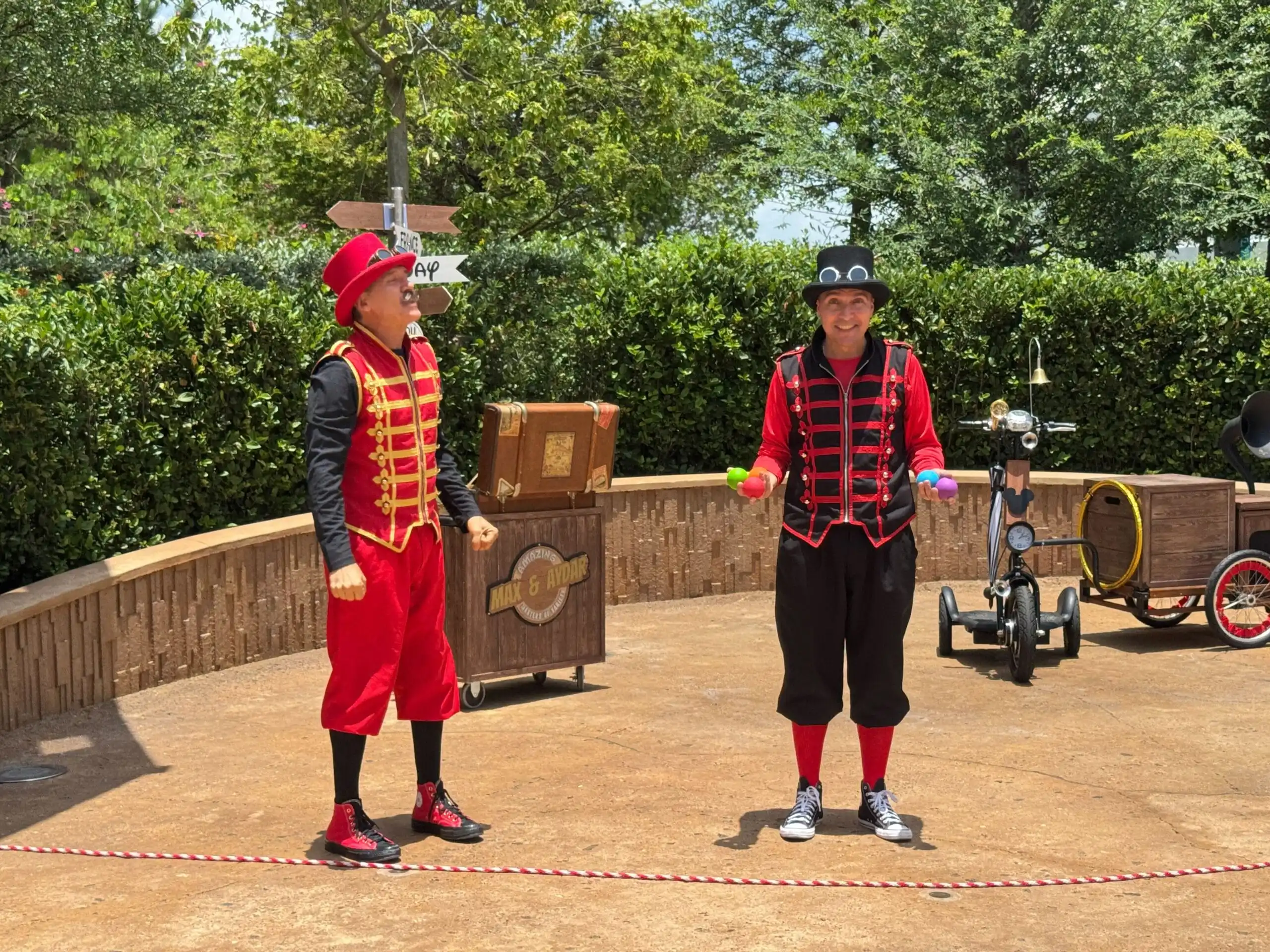 Two street performers in red and black costumes provide live entertainment outdoors near props, a bicycle, and EPCOT.