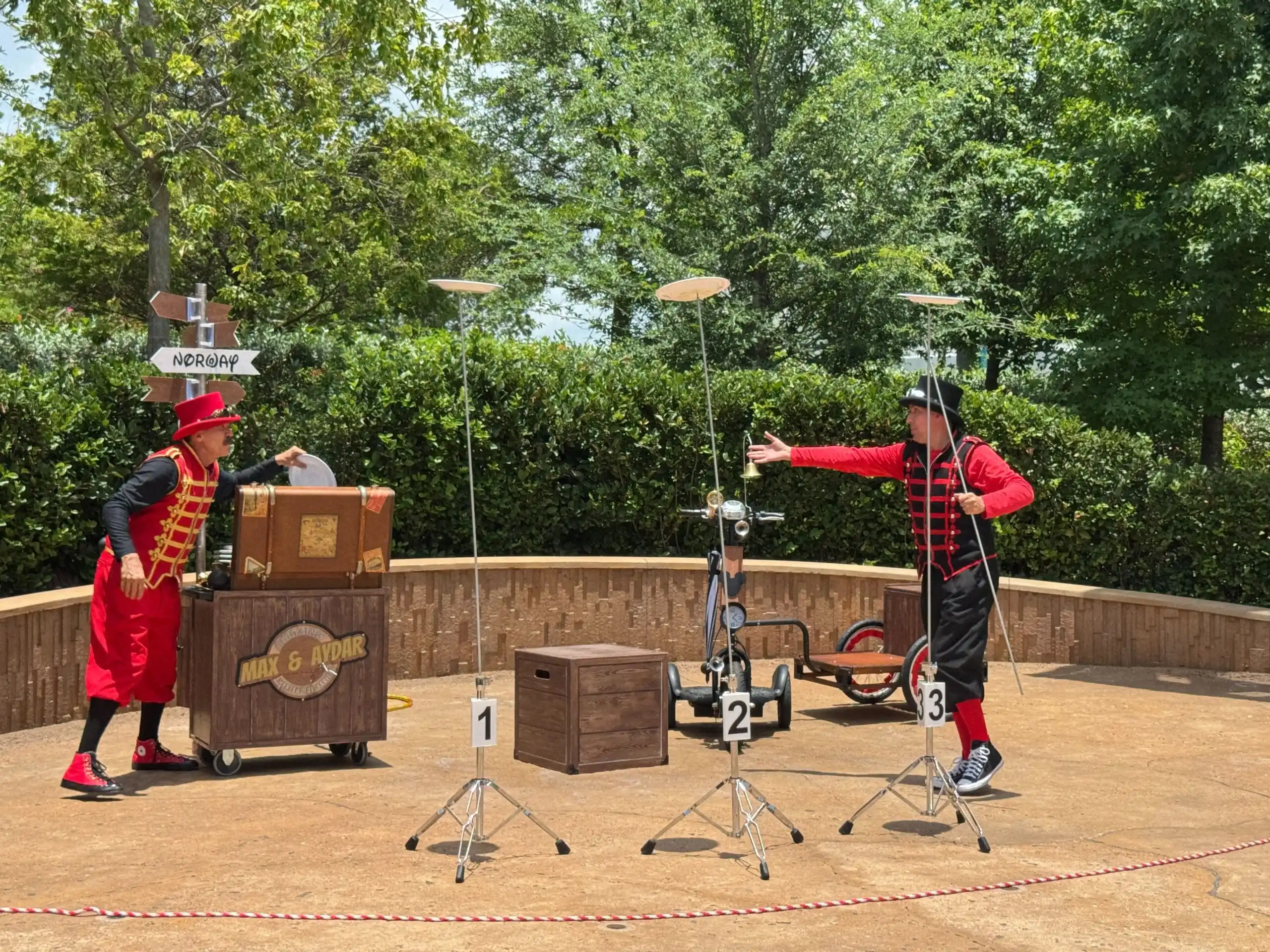 Two performers in colorful costumes spin plates on poles at an EPCOT outdoor entertainment show, surrounded by trees and props.