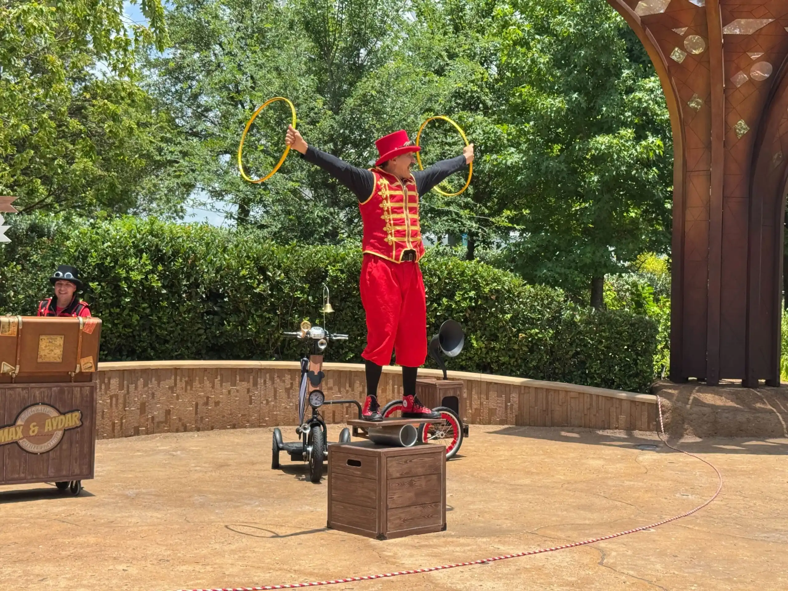 A circus performer in red stands on a wooden box with yellow hoops, bringing new entertainment to EPCOT on a sunny day.