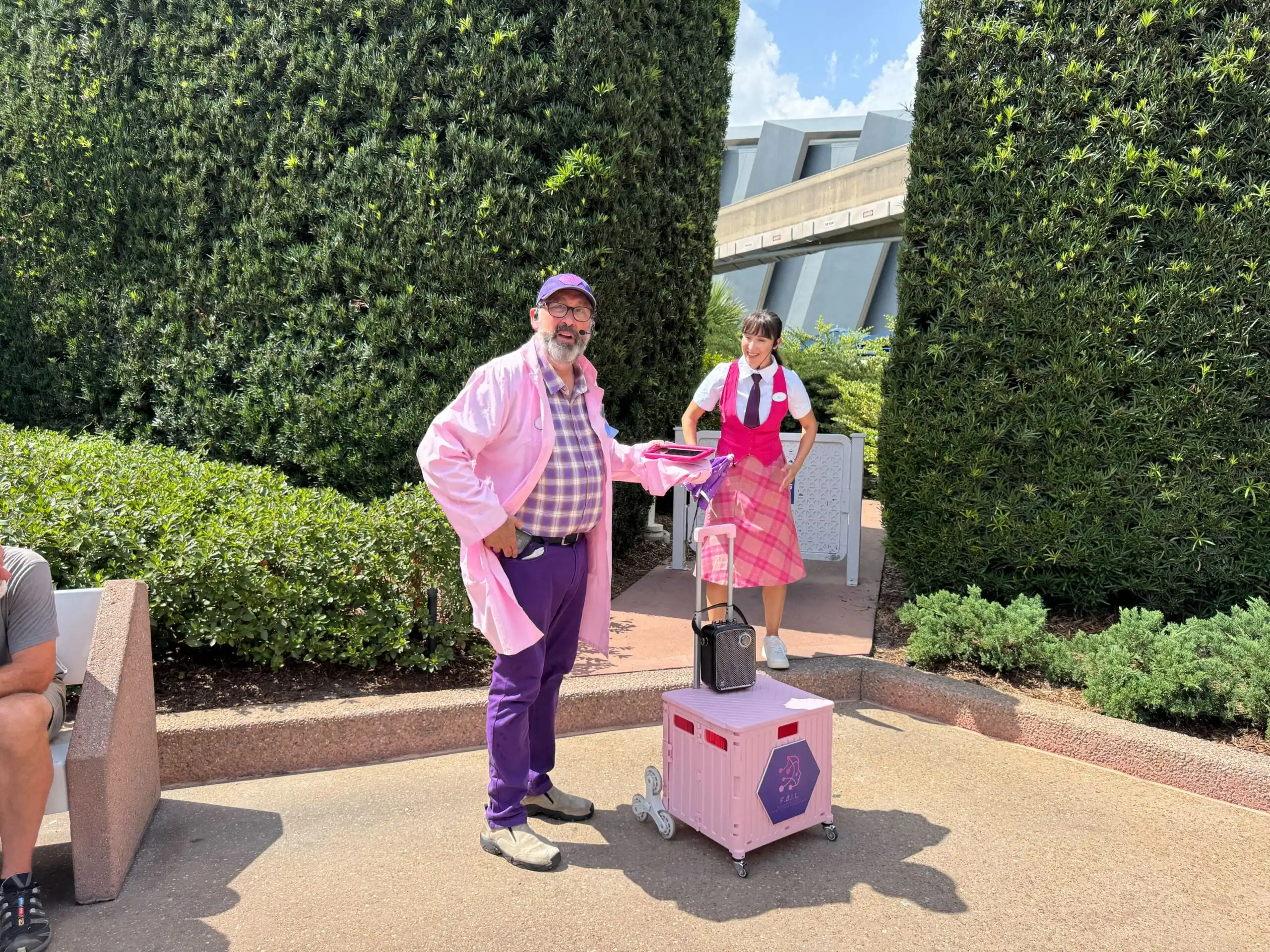Two people in pink costumes and a small pink cart create New Entertainment near tall hedges at EPCOT.