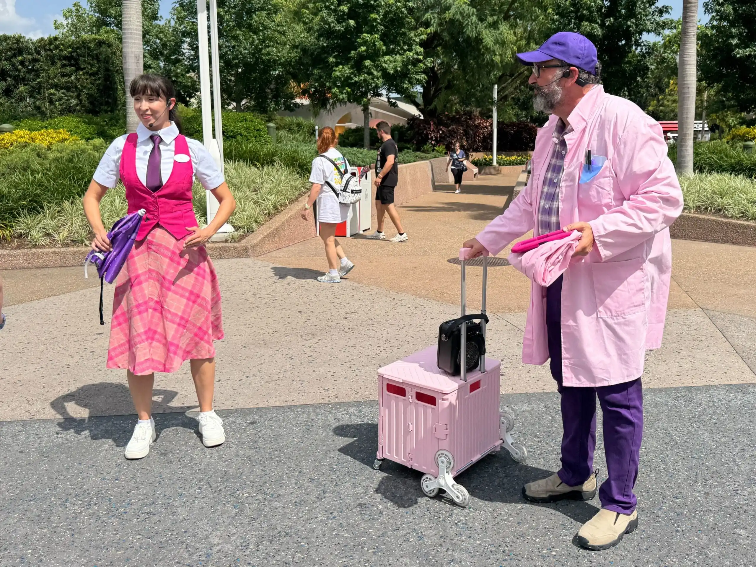 Two people in pink outfits stand outside EPCOT, one with a pink suitcase and the other with a purple bag.