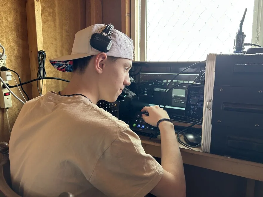 A high school boy sits in front of a table wearing a tan-colored tee shirt, a white baseball hat on backwards, and black headphones on top of his head. He's looking at a video screen that shows a camera angle of the baseball diamond. His hand is on top of a button control panel with numbers that controls which camera angle he wants to show. In front of him is another screen with more technical camera information on it. 
