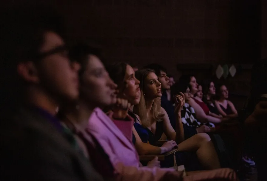 Students wearing fancy clothes look to the right. They are seated in a dark auditorium in a row of chairs. 
