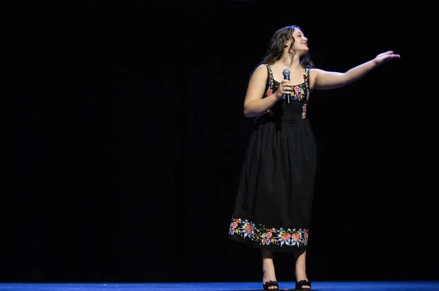 A girl with curly brown hair gestures to the right and smiles on a stage. She's wearing a black heels and a black dress embroidered with colorful, pink flowers on the straps, chest and edge of the skirt. She's also holding a microphone.