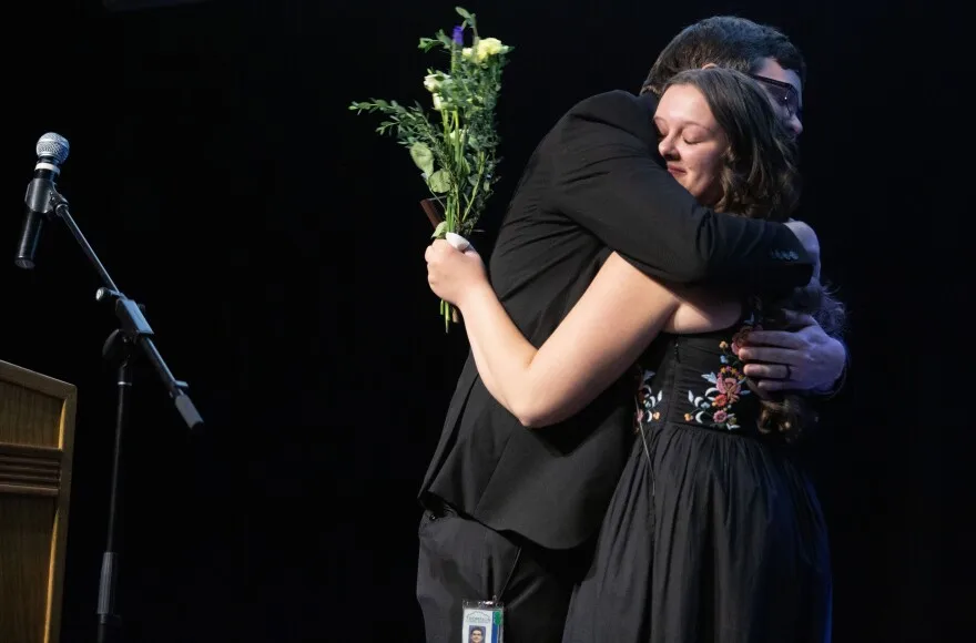 A man with brown hair and glasses wearing a black blazer and pants hugs a girl wearing a black dress with embroidered pink flowers on it. The girl is to the right of him. She is smiling and holding a bouquet of yellow flowers in her left hand. To the left of them is a wooden podium with a microphone on a mic stand.