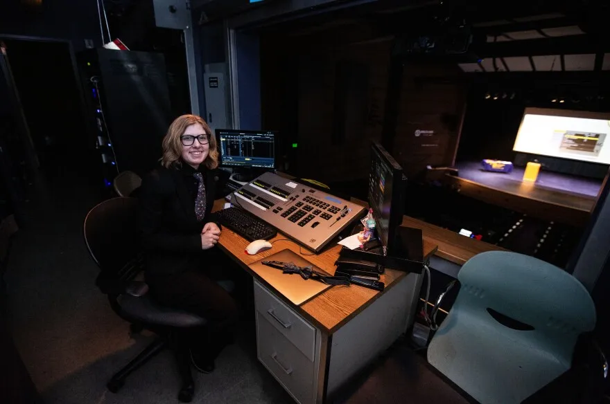 A girl with wavy, brown hair and glasses wearing a black button-up shirt and pants with a flowery tie smiles while sitting at an office chair. She is in front of a wooden desk which has a lighting control panel with several black buttons on it. To the left and right of the control panel are computer monitors. The desk faces a opening that looks out onto an auditorium with seats and a stage.