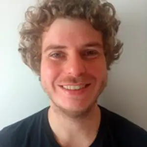 Jovem branco, cabelo castanho claro encaracolado, barba e bigode ralos e sorrindo para a câmera. Veste camiseta de cor preta
