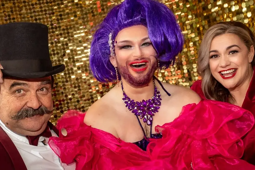 A man tipping his top hat stands with a drag queen in pink ruffles dress and purple hair next to a woman in red blazer