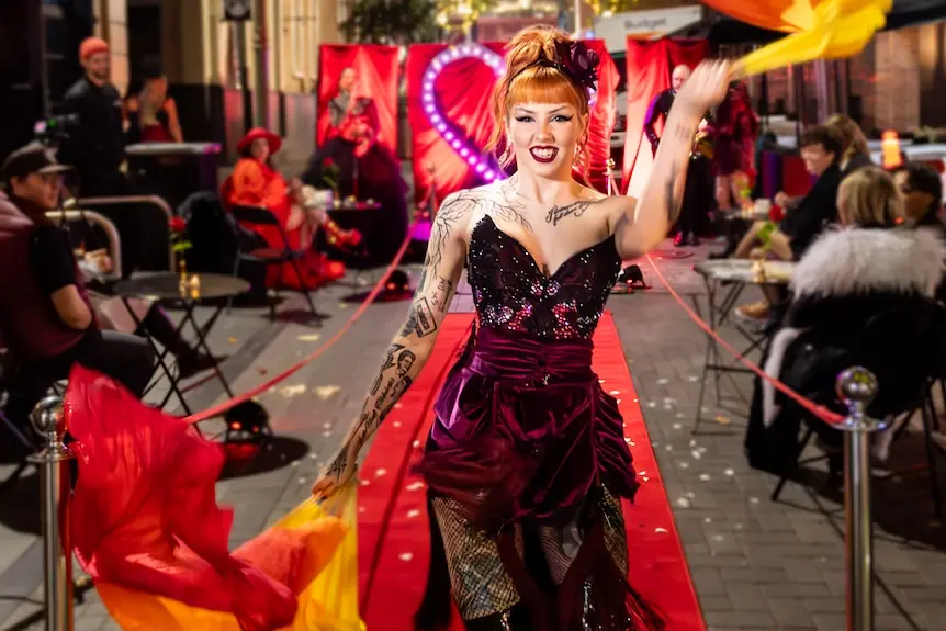 A performer in a corset dress stands at the end of a red carpet waving a bright scarf