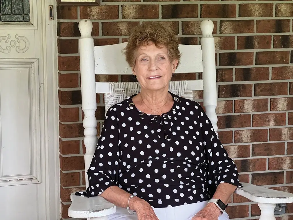 Old woman in a black shirt with white polka dots sitting in a white rocking chair on a porch