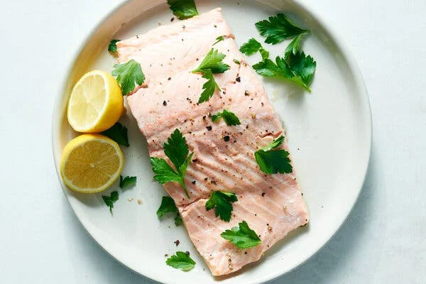 A white plate holds a pink fillet of microwave salmon garnished with parsley leaves and lemon halves.