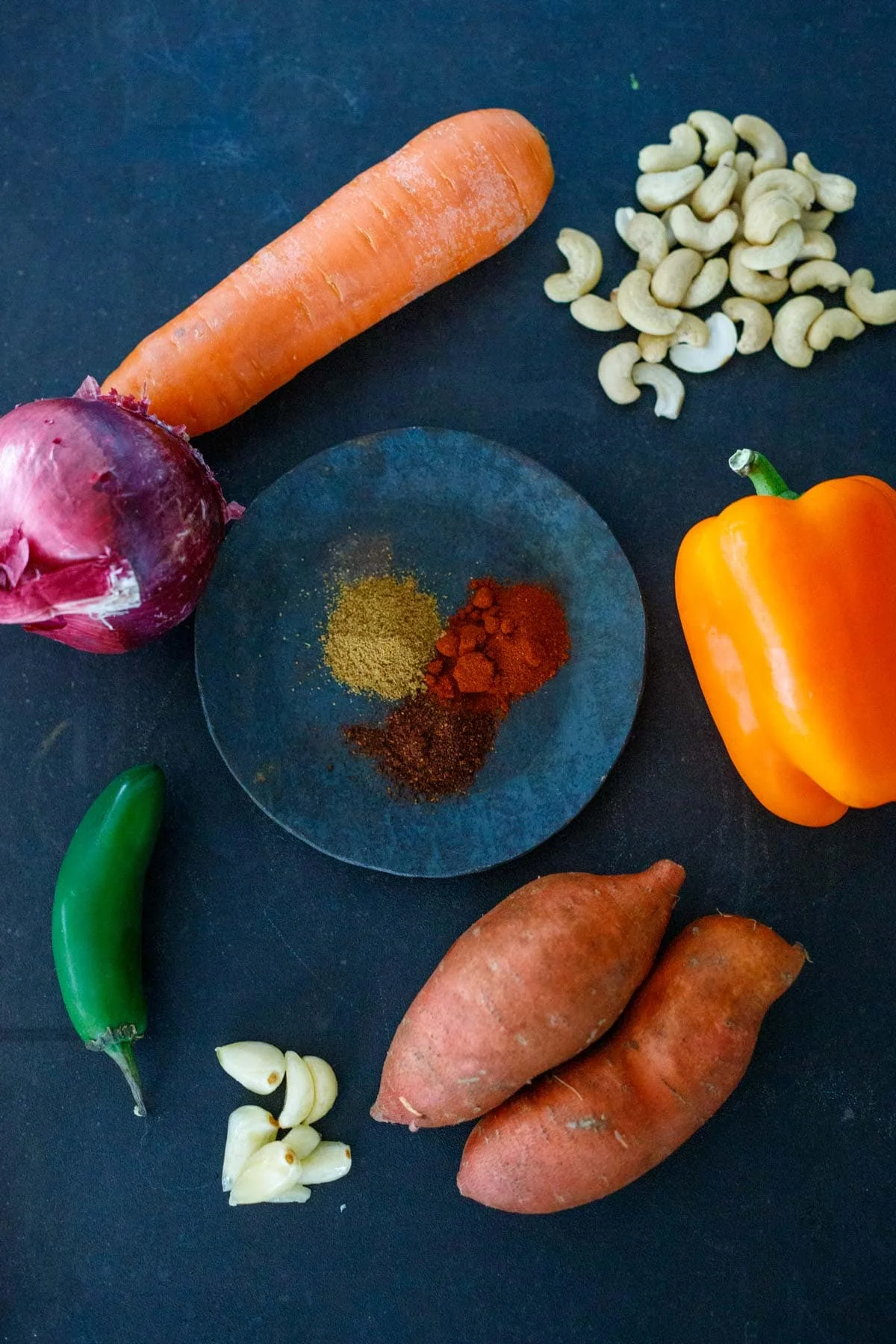 ingredients in vegan queso- spices, cashews, garlic cloves, orange bell pepper, carrot, sweet potatoes, red onion.