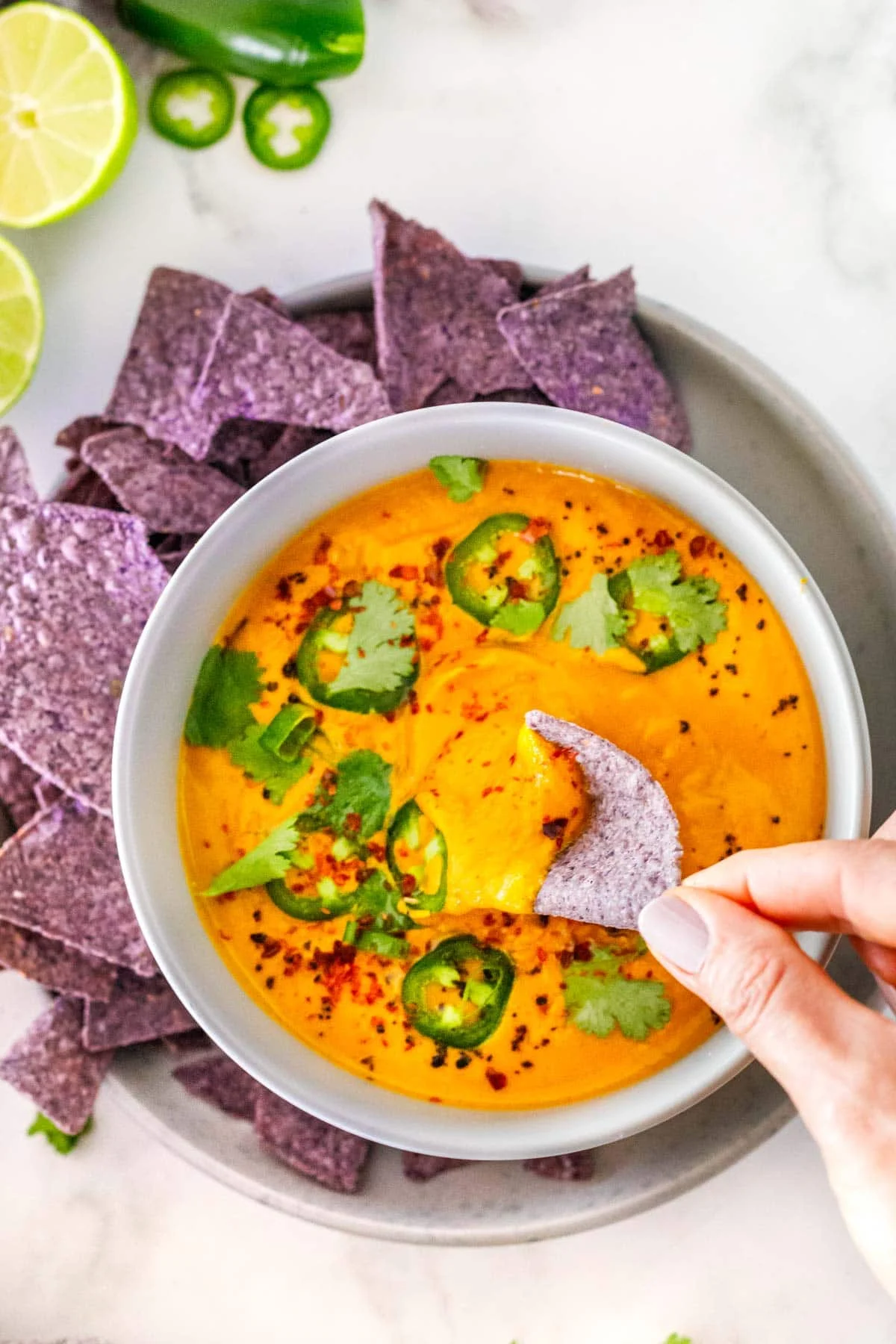vegan queso dip in a bowl with jalapeños, scallions, cilantro, and red pepper flakes and a hand dipping a tortilla chips in.