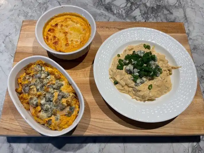 Straight-on shot of a cutting board with three bowls and plates of Buffalo-chicken dips. One bowl has a dip with crumbled blue cheese on top, one bowl has a dip with melted yellow cheese, and one dip is topped with crumbled blue cheese and scallions