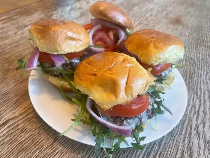 A white plate of four burgers with arugula, tomato slices, red onions, and brioche bun on a table