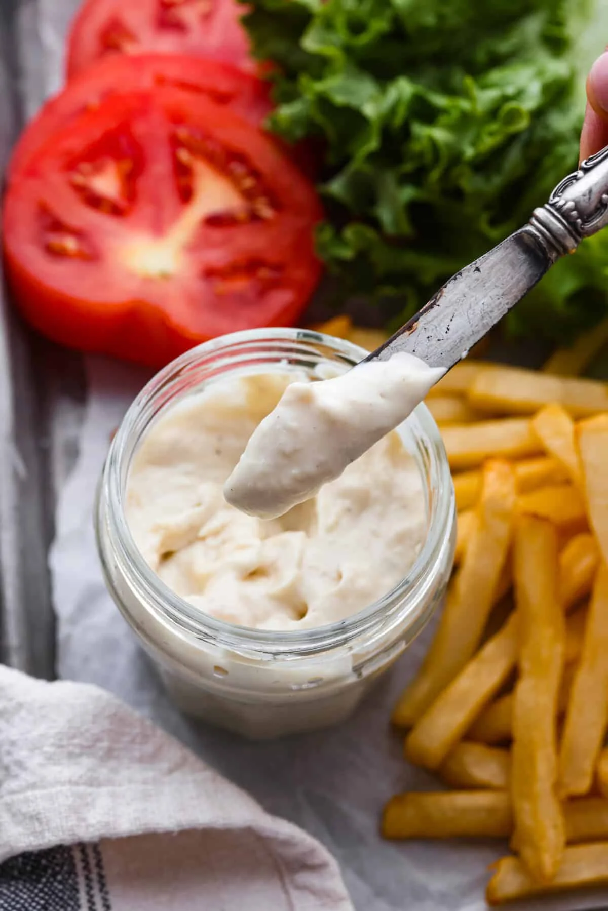 Close top view of a knife lifting donkey sauce from the jar. Fries and burger ingredients are scattered around the jar.