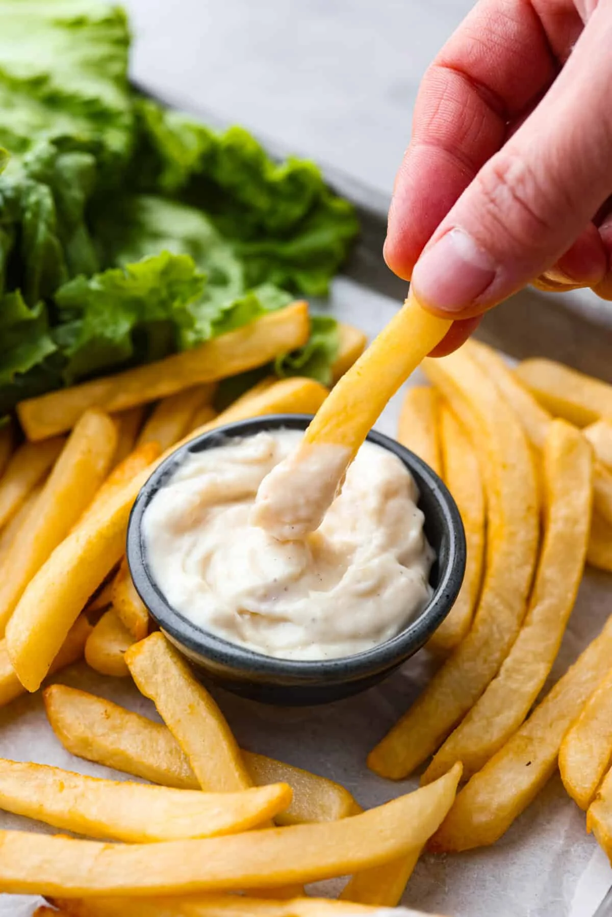 Close view of a French fry dipping in a bowl of donkey sauce. French fries and burger ingredients are scattered around the jar.