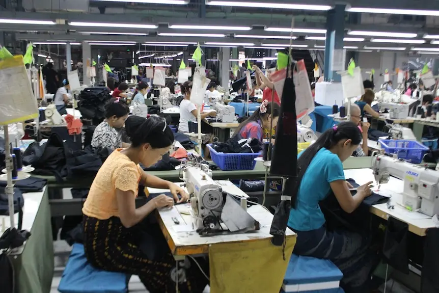 Two women huddle over sewing machines as they sew the hems of garments.