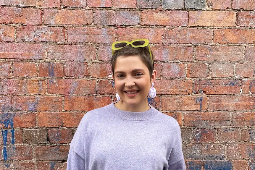 A young woman with short cropped hair and pastel purple knit jumper in front of a red brick exterior wall.