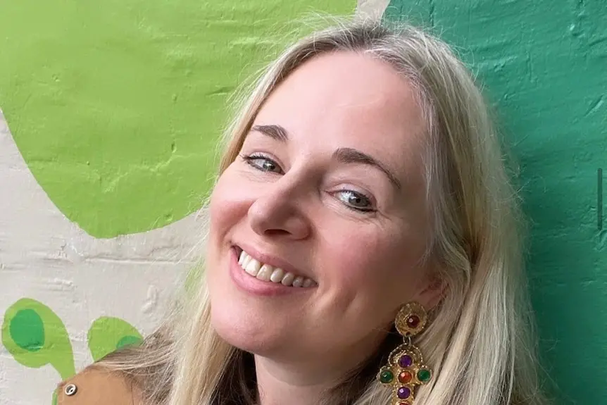 A woman with long blonde hair smiling with teeth in front of a colourful painted outside exterior wall.