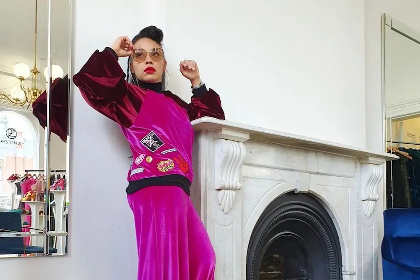 A woman in pink velvet flared pants and a red and pink velvet top posing against a fireplace.