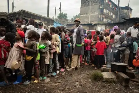 A line of ragged African children holding plates in front of tin shacks