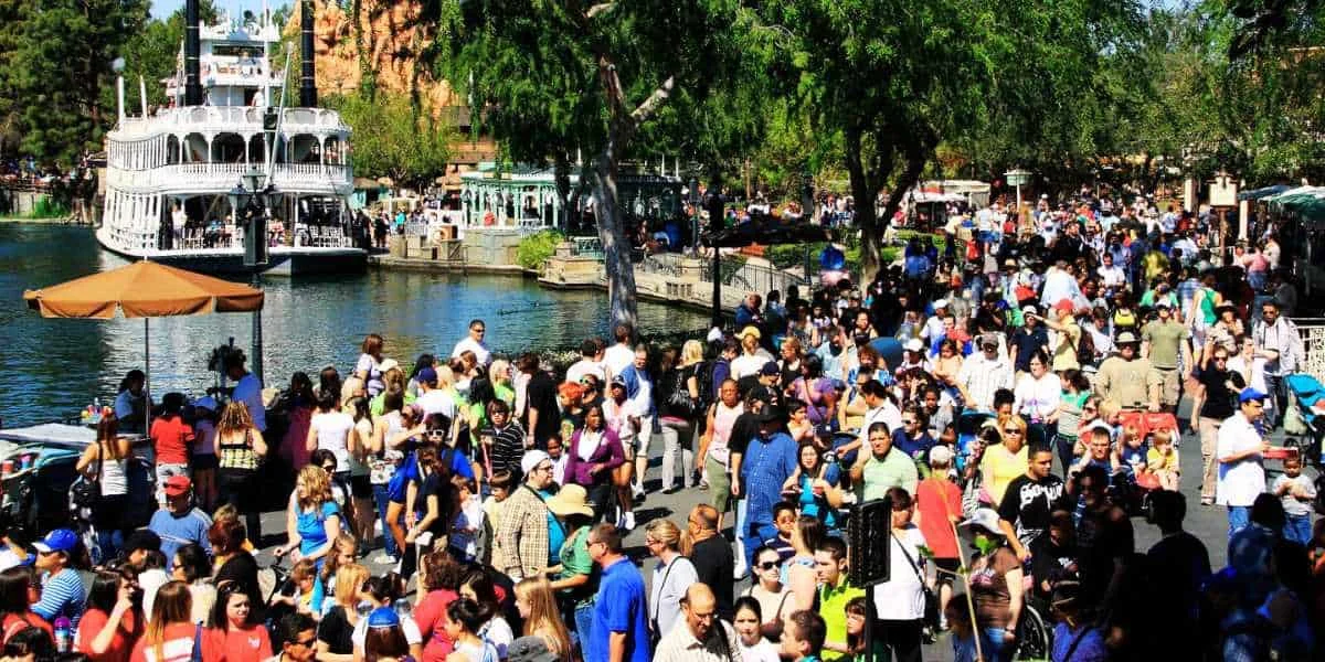 A busy outdoor scene at a Disney theme park with a large crowd of people, Disney merchandise stalls, a riverboat in the background, trees, and a clear blue sky.