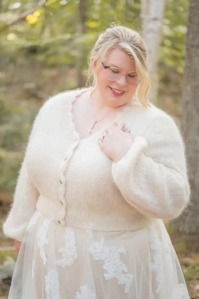 A smiling woman wearing a cream cardigan over a cream wedding dress as she lowers her gaze dreamily.