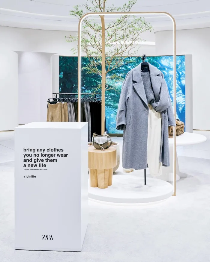 A modern and minimalistic Zara store setup showcasing a light gray coat and accessories on a wooden display stand. A prominent sign invites shoppers to recycle their old clothes through the #joinlife program