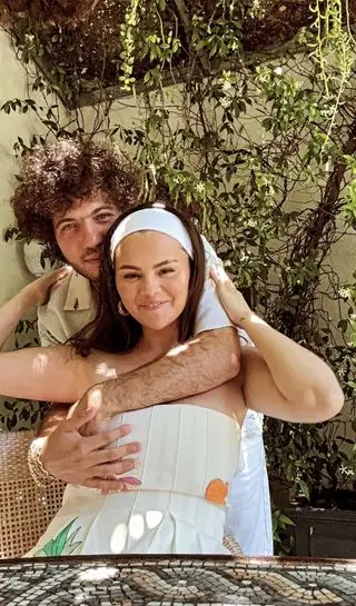 Selena Gomez and Benny Blanco on an outdoor patio where Selena Gomez wears a white strapless free people maxi dress and matching white headband