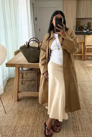 a photo of a summer rainy day outfit with a woman wearing a tan trench coat over a white t-shirt and off-white satin slip skirt with brown flip flops and a woven tote bag