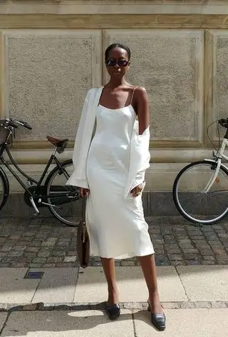 a photo of a summer rainy day outfit with a woman wearing a white button-down shirt over a white slip dress with black mules, black sunglasses, and a shoulder bag