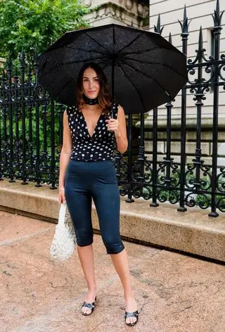 a photo of a summer rainy day outfit with a woman wearing a black umbrella, polka dot blouse, black capri pants, black heeled mules, and a white woven Bottega Veneta Bag