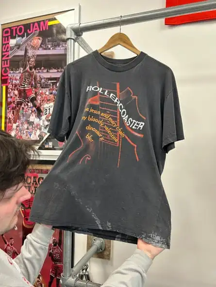 A man lifts the edge of a distressedT-shirt labelled “Rollercoaster, the Jesus and Mary chain, bloody valentine, dinosaur Jr, blur” T-shirt with curved bleach marks that’s on a hanger behind a basketball poster