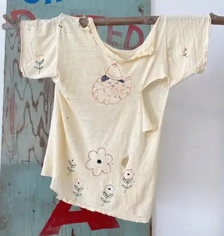 A moth-eaten T-shirt made of very worn cotton with a lightly embroidered clown’s face and flowers and rips at the collar and under a short sleeve