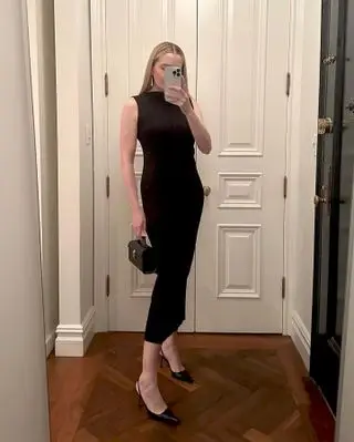 Who What Wear editor Kristen Nichols poses for a mirror selfie in a New York City apartment with herringbone wood floors wearing a boatneck midi black dress, mini Savette bag, and black slingback heel pumps