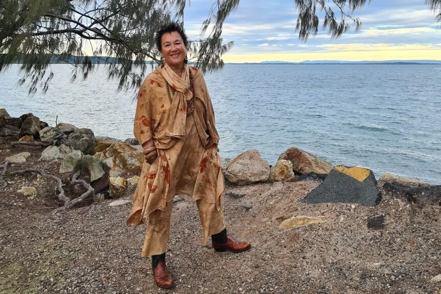 A woman stands on the banks of a lake wearing a long brown dress smiling at the camera.