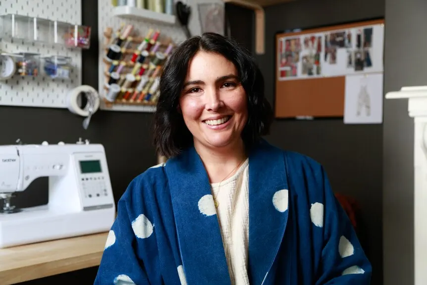 A woman smiles at the camera. In the background is a sewing machine and a wall of coloured thread.