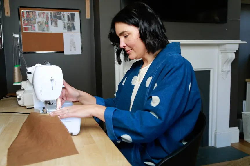 A woman sits at a table feeding a piece of fabric through a sewing machine.